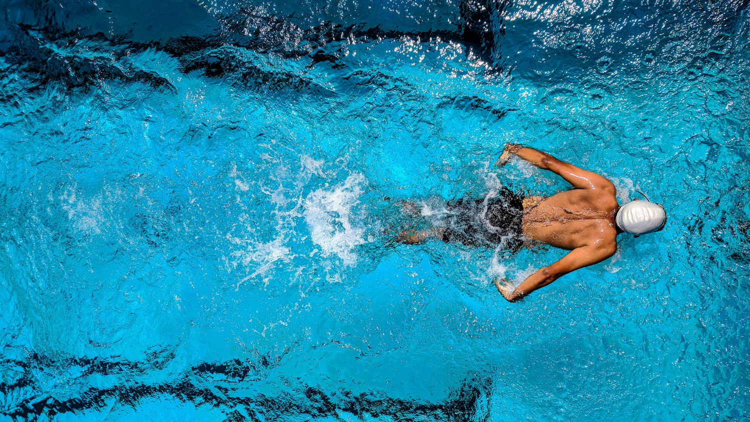 Top-down view of a male swimmer performing freestyle in a clear blue pool, creating ripples and splashes