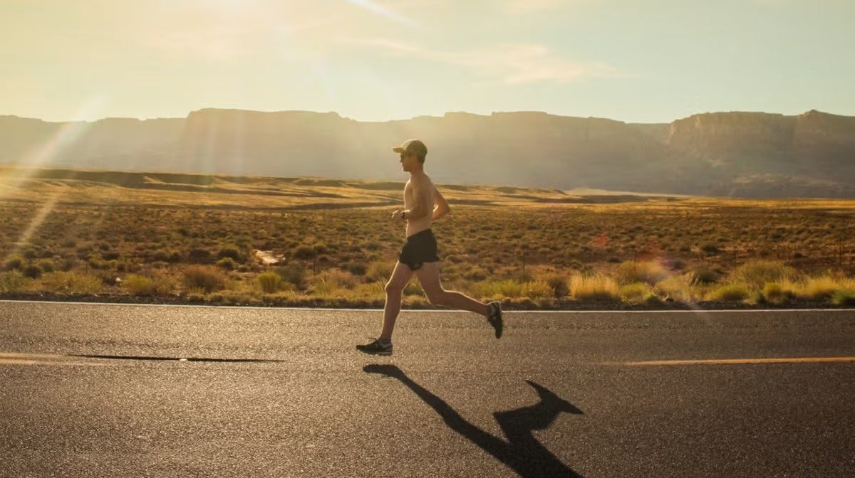 Man running shirtless on a desert highway at sunrise, with mountains in the background and long shadows on the road